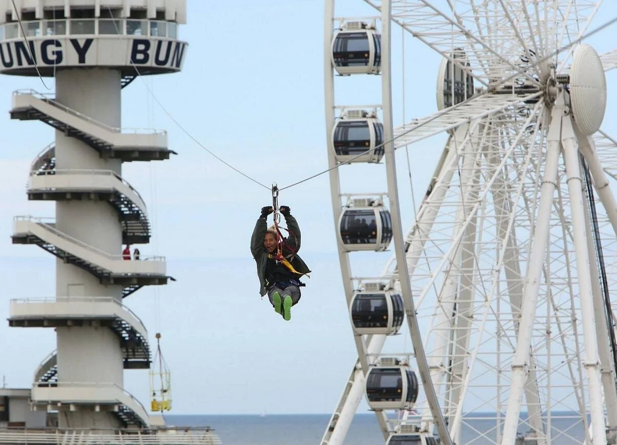 Visitor riding the Scheveningen zipline with SkyView Ferris wheel and Bungy Tower behind